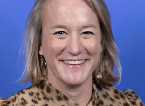 A person with shoulder-length blonde hair, wearing a leopard print blouse, smiling against a blue background.