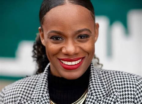 A person smiling, wearing a black and white checkered blazer, a black top, and a gold necklace, with a blurred green background.