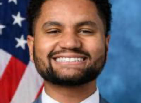 Smiling man in a suit and tie with an American flag in the background.