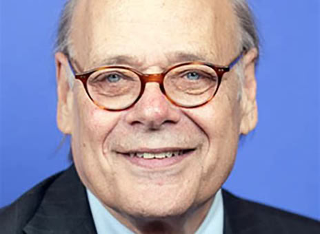 A man with glasses, wearing a suit and an orange tie, against a blue background.