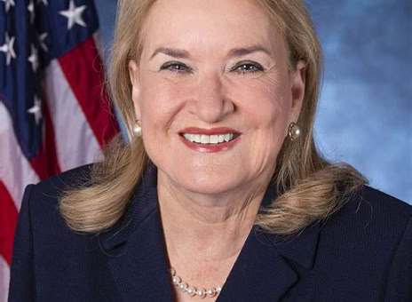 A woman with blonde hair wearing a dark suit and pearl necklace, smiling in front of an American flag and blue background.