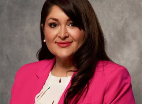 A person with long dark hair wearing a bright pink blazer and a white blouse, set against a gray background.