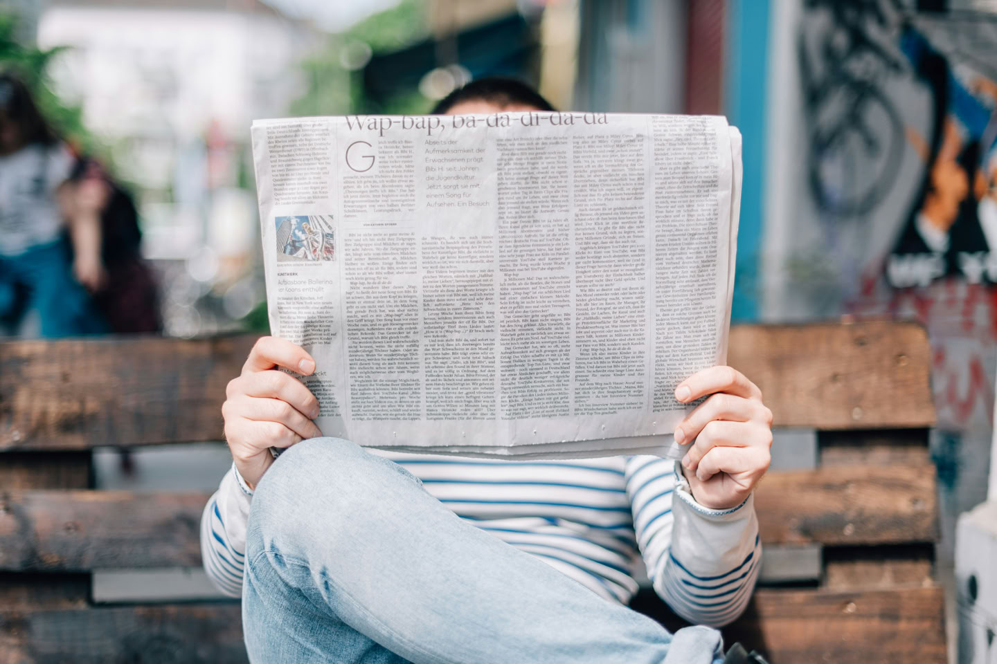 Person sitting on a bench reading a newspaper, with legs crossed and face obscured by the newspaper.