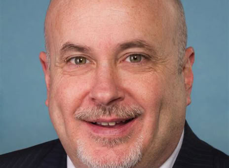 A bald man with a goatee, wearing a dark suit, white shirt, and blue striped tie, smiling against a plain blue background.