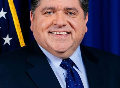 A man in a suit and tie smiling in front of a blue curtain with part of a flag visible.