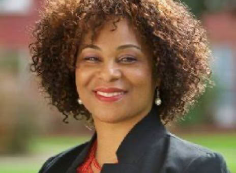 A person with curly hair wearing a black blazer and red top, smiling outdoors with a blurred background.