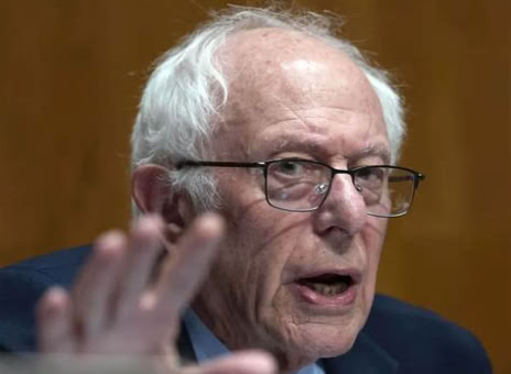 Elderly man with white hair and glasses speaking, hand raised in foreground.