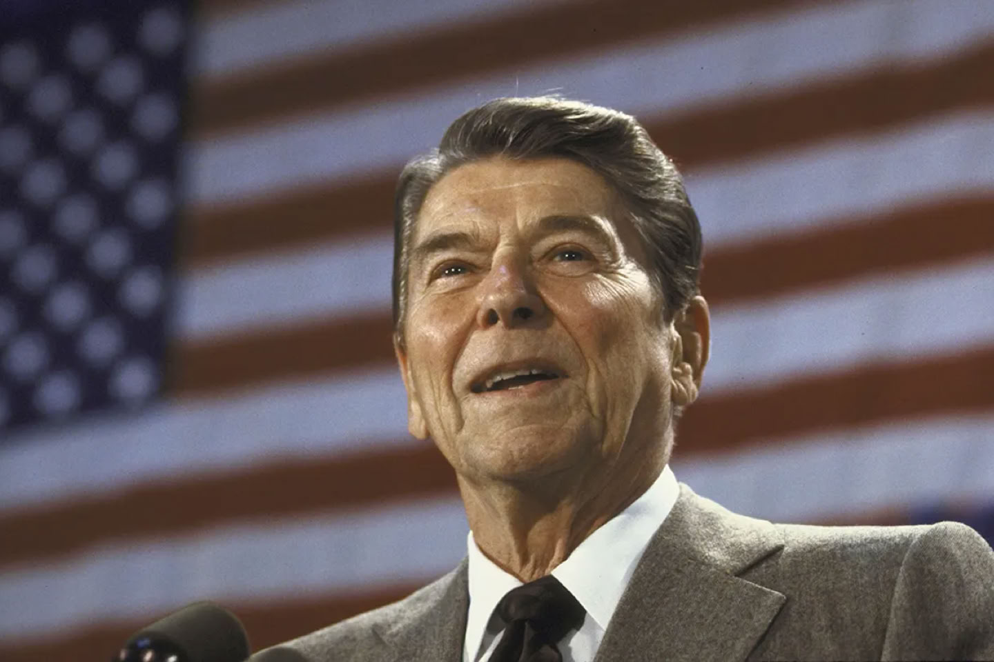 President Ronald Reagan in a suit speaking in front of a large American flag.