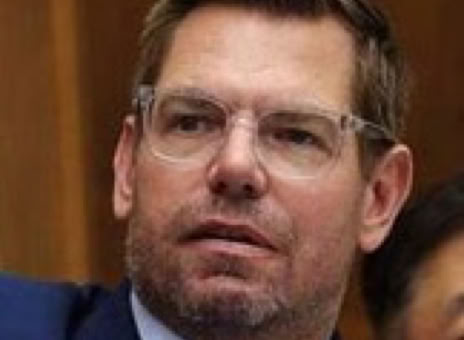 Man with glasses and a beard wearing a suit and tie, sitting indoors.